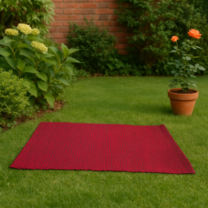 "Red woven yoga mat placed on a green garden lawn with flowering plants and a potted rose, creating a peaceful outdoor setting."